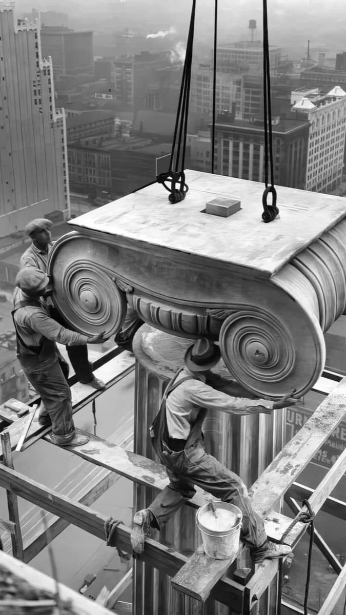 Construction workers guiding an ionic column capital into place atop a skyscraper